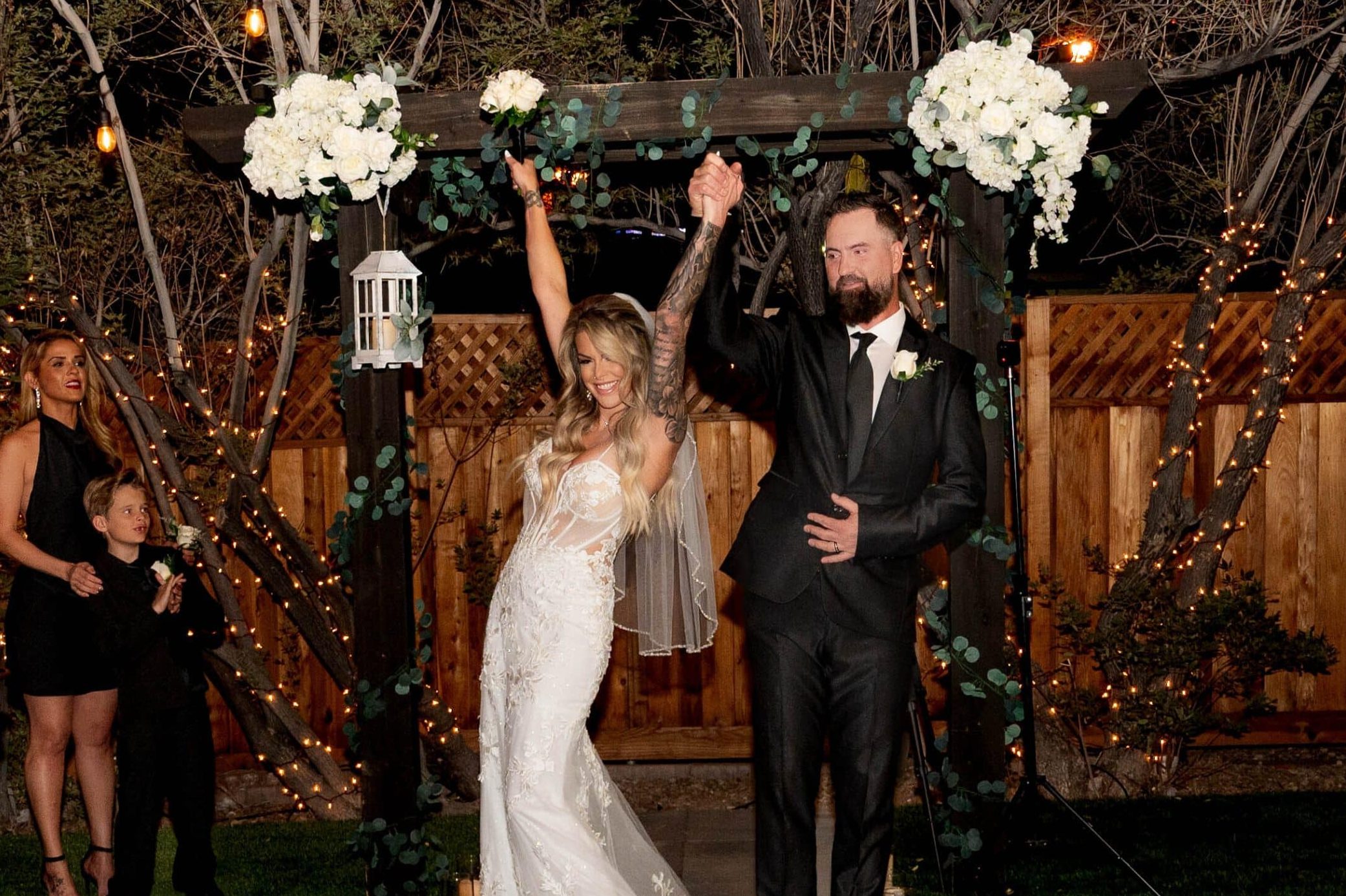 Couple holding hands under a canopy of twinkle lights surrounded by greenery and fresh floral arrangements at Mon Bel Ami Wedding Chapel.